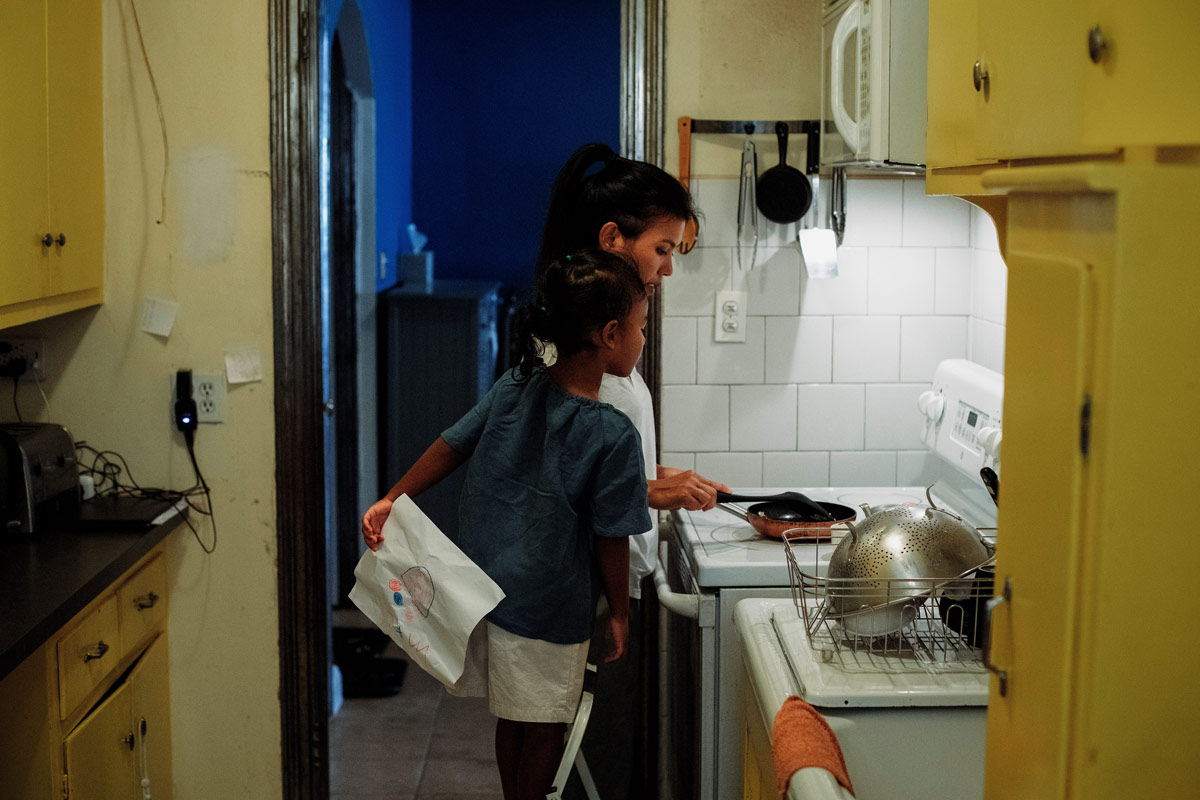 An adult cooking on an electric stovetop in the kitchen, with a child standing on a step stool on the right of the adult, watching them cook